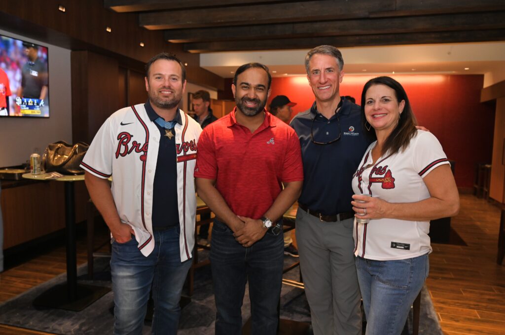 An Evening of Honor: Celebrating Leadership and Service 1 Four people pose together in a suite at a baseball game, smiling at the camera. One wears a Medal of Honor and Braves jersey, and another sports a red Braves polo. A game plays in the background.